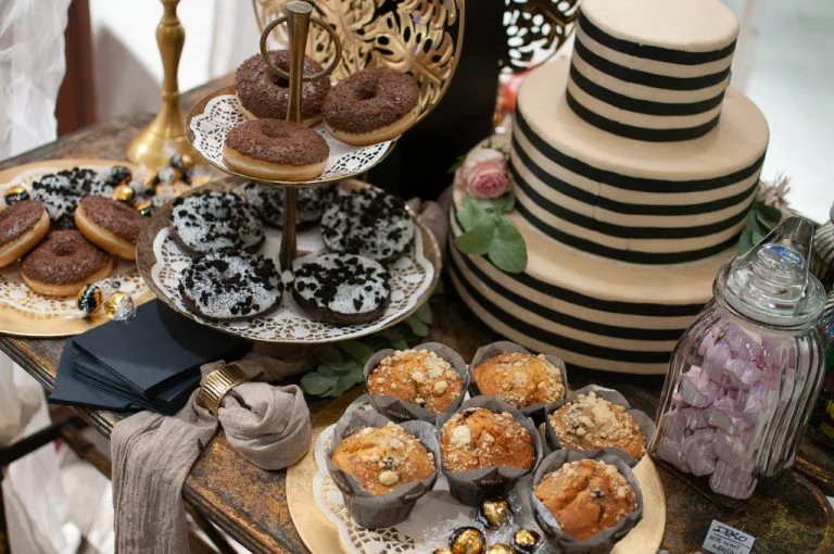 Ein verlockender Blick auf einen süßen Tisch auf der "Trau - Die Hochzeitsmesse"präsentiert eine Auswahl an Köstlichkeiten. Im Vordergrund sind saftige Muffins in Papierförmchen auf einer goldenen Platte angerichtet, umgeben von glänzenden Goldpralinen. Dahinter steht eine elegante, zweistöckige Hochzeitstorte in Beige mit breiten, schwarzen Querstreifen und zarten rosa Blüten als Dekoration. Auf einer goldenen Etagere werden Schokoladen-Donuts und Donuts mit weißem Zuckerguss und schwarzen Streuseln präsentiert. Der Tisch ist zudem mit weiteren Süßigkeiten und Servietten dekoriert. Die Szene vermittelt einen Eindruck von den vielfältigen süßen Optionen, die für eine Hochzeitsfeier angeboten werden.