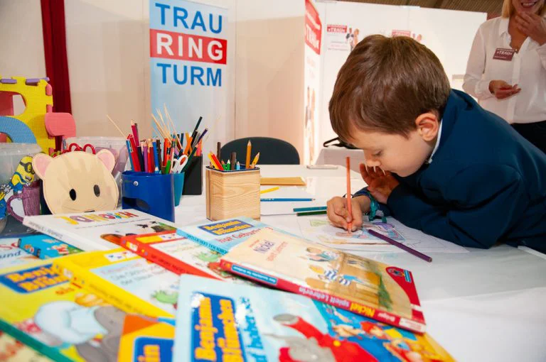 Ein kleiner Junge vertieft sich in ein Malbuch an einem Kindertisch auf der "Trau - Die Hochzeitsmesse ". Der Tisch ist mit Malbüchern, Buntstiften in verschiedenen Behältern und anderen Spielsachen gefüllt. Im Hintergrund ist ein Banner mit der Aufschrift "TRAURINGTURM" zu erkennen, was darauf hindeutet, dass dieser Bereich möglicherweise eine Kinderbetreuung oder eine Spielecke für die kleinen Besucher der Hochzeitsmesse ist. Eine erwachsene Person steht im Hintergrund und beobachtet das Kind. Die Szene zeigt, dass auch an die Unterhaltung der jüngsten Gäste gedacht wird, während ihre Eltern die Messe erkunden.