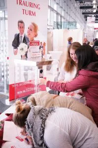 Drei junge Frauen beugen sich über einen Tisch am Stand von "TRAURING TURM" auf der "Trau - Die Hochzeitsmesse "und scheinen sich für die ausgestellten Trauringe zu interessieren. Ein großer Banner im Hintergrund zeigt ein glückliches Brautpaar und wirbt mit "Tipps. Modenschauen. Trends." sowie der Website-Adresse. In einer beleuchteten Vitrine auf dem Tisch werden verschiedene Eheringe präsentiert. Die Frauen wirken interessiert und vertieft in ihre Auswahl. Die Atmosphäre am Stand ist geschäftig und informativ für Paare, die auf der Suche nach ihren perfekten Trauringen sind.