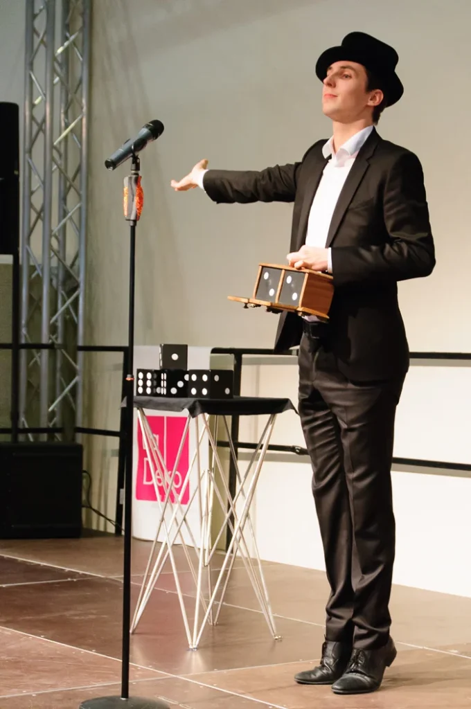 Ein junger Magier in einem eleganten schwarzen Anzug und einem schwarzen Hut präsentiert auf einer Bühne der "Trau - Die Hochzeitsmesse " einen Zaubertrick mit Würfeln und einer kleinen Holzkiste. Er steht selbstbewusst vor einem Mikrofonständer und gestikuliert mit der einen Hand, während er in der anderen Hand die geöffnete Kiste hält, in der ein großer Würfel mit der Zahl Sechs nach oben zeigt. Auf einem kleinen Tisch neben ihm liegen weitere Würfel und magische Requisiten. Die Bühne ist schlicht gehalten mit einem hellen Hintergrund. Die Szene fängt einen spannenden Moment der Unterhaltung ein, der die Besucher der Hochzeitsmesse in Staunen versetzt.