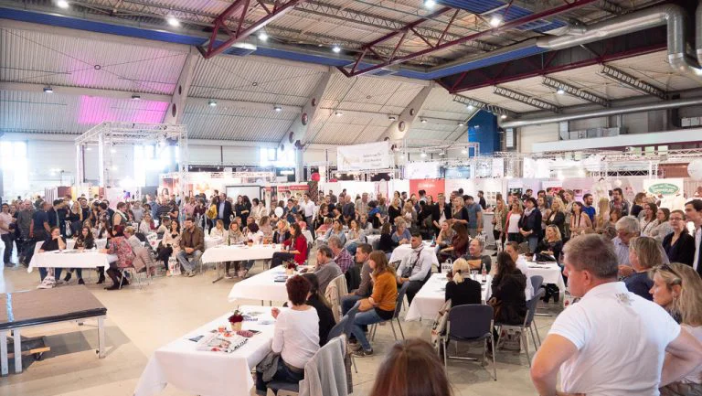 Ein belebter Moment auf der "Trau - Die Hochzeitsmesse " zeigt eine große Menschenmenge, die an Tischen sitzt und aufmerksam einer Präsentation oder Modenschau folgt. Die Halle ist hell erleuchtet und mit zahlreichen Messeständen im Hintergrund gefüllt. Die Besucher scheinen gespannt auf das Programm zu sein. Die Atmosphäre ist erwartungsvoll und geschäftig, typisch für eine gut besuchte Hochzeitsmesse.