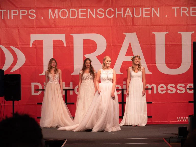 Vier junge Frauen präsentieren auf einer Bühne der "Trau - Die Hochzeitsmesse 2026" elegante Brautkleider. Die Kleider sind in verschiedenen Weiß- und Cremetönen gehalten und zeigen unterschiedliche Schnitte und Verzierungen, von zarter Spitze bis hin zu fließenden Stoffen. Drei der Models tragen Blumenkränze im Haar. Im Hintergrund ist ein großer, roter Banner mit dem Wort "TRAU" und dem Schriftzug "Die Hochzeitsmesse" zu sehen, umrahmt von den Begriffen "TIPPS", "MODENSCHAUEN" und "TRENDS". Die Szene fängt einen Moment der Brautmodenschau ein, bei dem die neuesten Trends der Saison präsentiert werden.